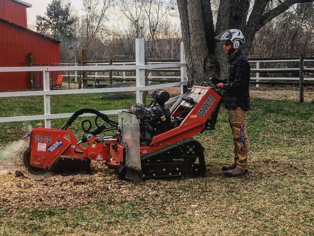 A Tapson&rsquo;s operator in a helmet and safety glasses working a tracked stump grinder at the base of a large tree, the cutting wheel throwing a spray of chips across the grass beside a white rail fence.